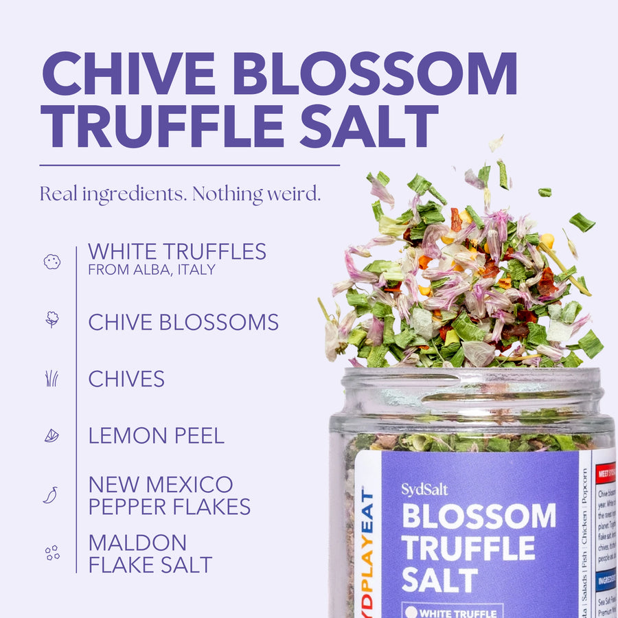 SydSalt Chive Blossom Truffle Salt in a clear glass jar with a purple label, shown next to a pile of chopped white truffles, chive blossoms, chives, lemon peel, pepper flakes, and flake salt on a light lavender background with ingredient callouts.