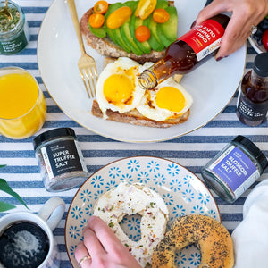 A colorful breakfast setup featuring avocado toast topped with cherry tomatoes and two fried eggs on toast, drizzled with chili truffle olive oil. A hand is pouring the oil, while Super Truffle Salt and Blossom Truffle Salt jars are placed beside the plates. A glass of orange juice, coffee cup, and fresh flowers complete the vibrant meal.