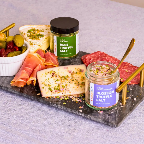 A close-up of the charcuterie board from Image 3, showcasing the Herb Truffle Salt and Blossom Truffle Salt jars, along with meats, cheeses, and olives. Gold utensils are visible, and the ingredients are arranged on a dark stone surface.