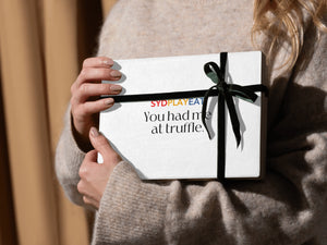 A woman in a cozy sweater holds a luxurious white SydPlayEat corporate gift box wrapped in a black ribbon, featuring the tagline 'You had me at truffle.' A perfect corporate gifting option for clients and employees.