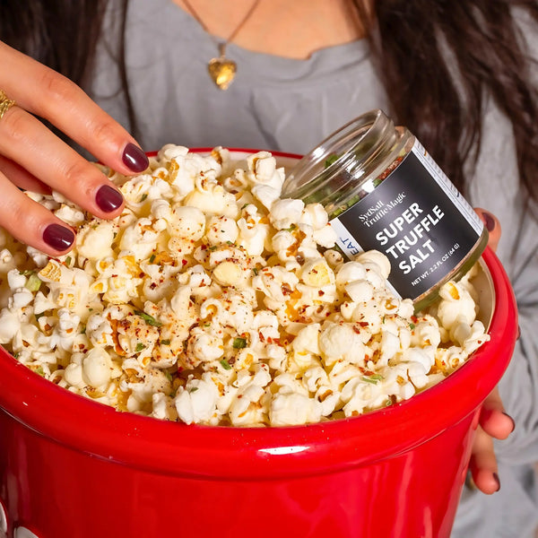 A close-up of a red bowl filled with popcorn, seasoned with SydPlayEat's Super Truffle Salt. A woman's hand is holding the salt jar as she sprinkles it over the popcorn, with the seasoning clearly visible on the popcorn.