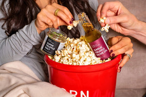 Two people, presumably a couple, enjoying a shared popcorn moment. One is adding Super Truffle Salt, while the other is pouring Garlic Truffle Olive Oil over the popcorn in a large red popcorn bowl.