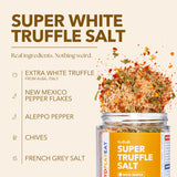 SydSalt Super White Truffle Salt in a clear glass jar with a yellow label, shown with a dramatic splash of the seasoned salt mixture. The background is a solid light beige with text detailing the ingredients: extra white truffle from Alba, Italy; New Mexico pepper flakes; Aleppo pepper; chives; and French grey salt.