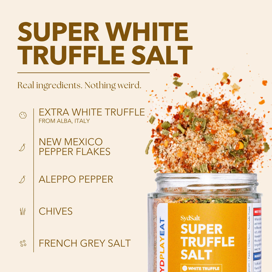 SydSalt Super White Truffle Salt in a clear glass jar with a yellow label, shown with a dramatic splash of the seasoned salt mixture. The background is a solid light beige with text detailing the ingredients: extra white truffle from Alba, Italy; New Mexico pepper flakes; Aleppo pepper; chives; and French grey salt.